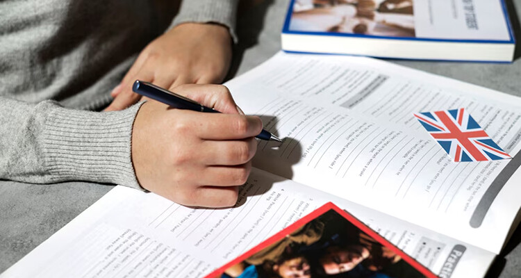 Close-up of a hand writing in an IELTS study book, with a UK flag, symbolizing preparation for IELTS booking